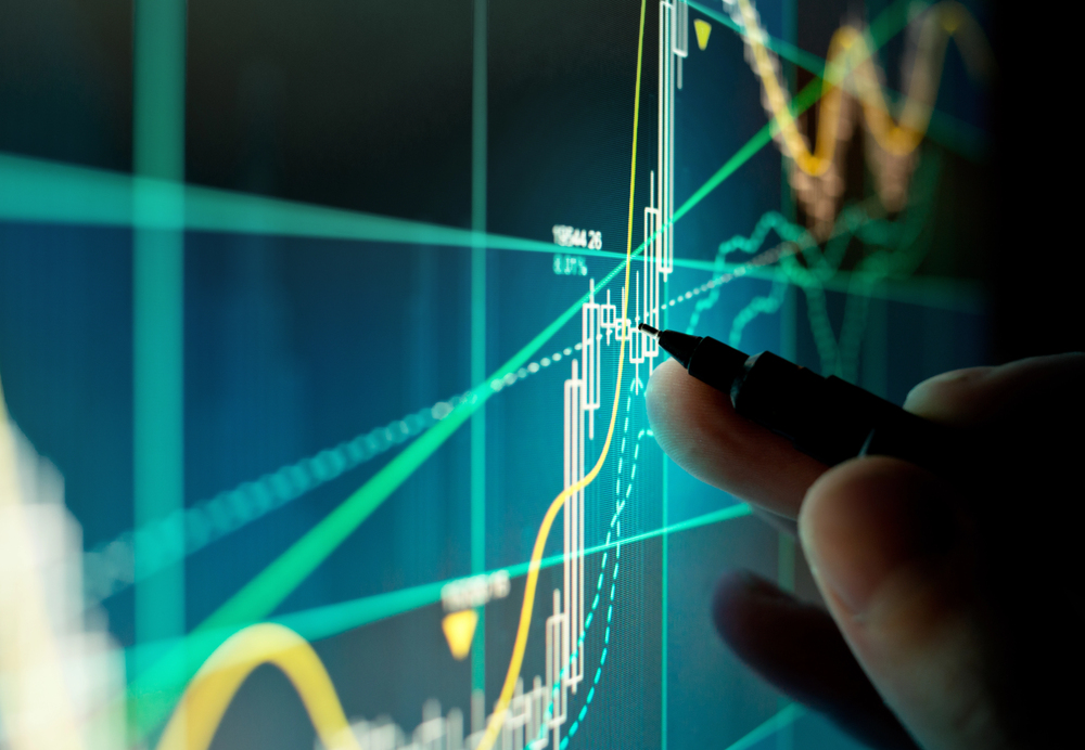 A hand holding a pen points to a digital financial chart on a screen, displaying various coloured lines and candlestick patterns indicating share market trends and analysis.
