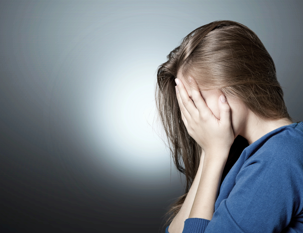 A woman with long brown hair, wearing a blue top, sits against a gradient grey background and covers her face with both hands, suggesting sadness or distress.