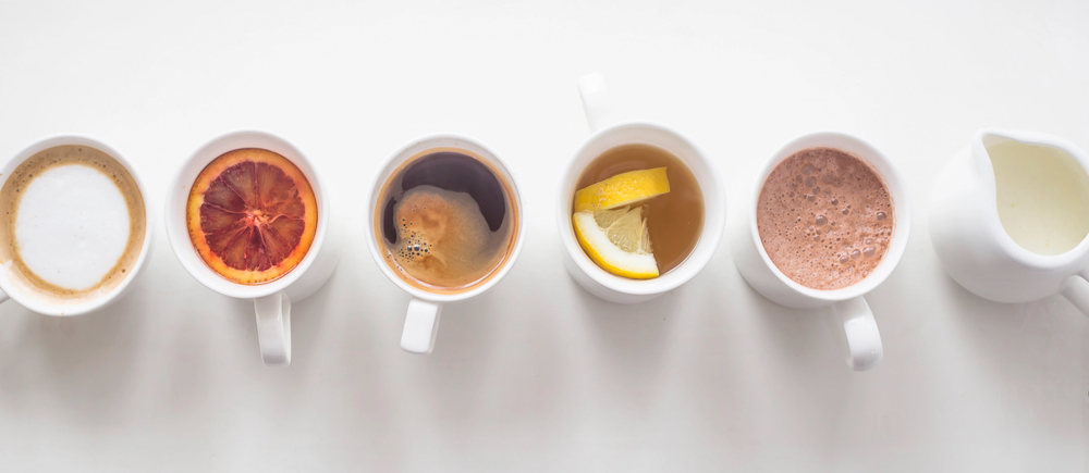 Six cups in a row on a white surface, each containing a different beverage: cappuccino, drink with a blood orange slice, black coffee, tea with lemon slices, hot chocolate, and a small jug of milk.