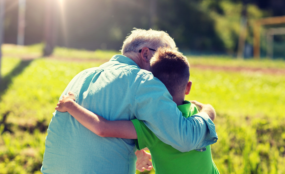 An older man and a young boy, seen from behind, sit outdoors in sunlight with their arms round each other, showing affection and closeness. The background is green and softly blurred.
