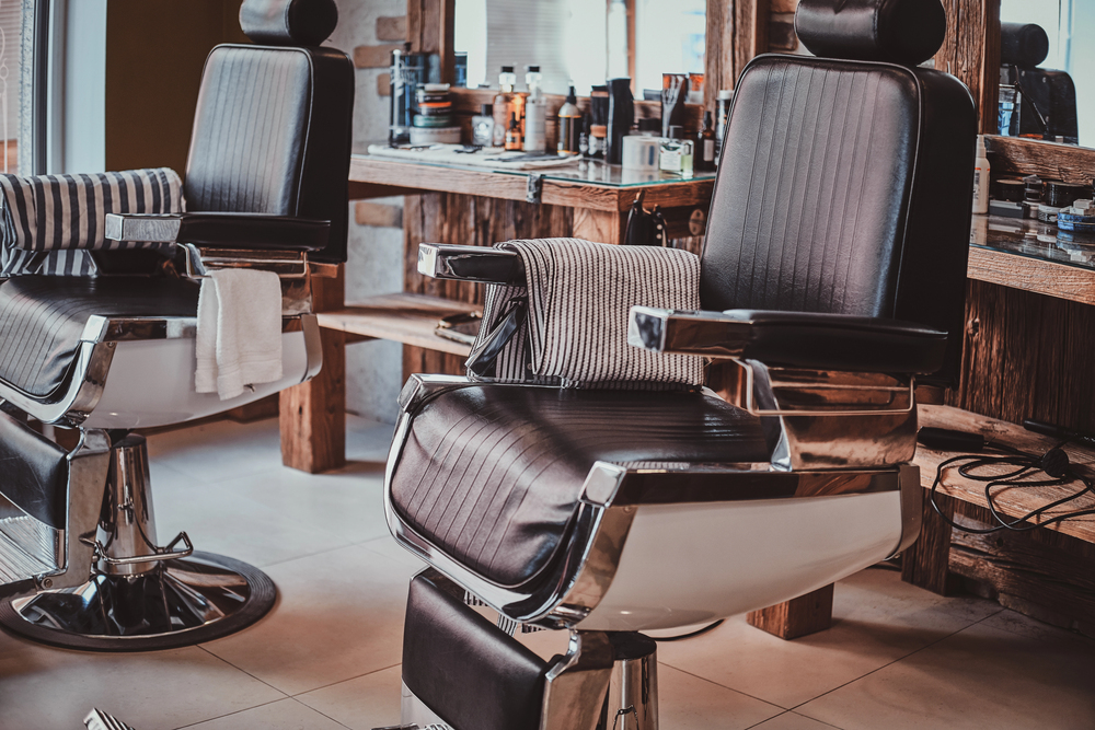 A stylish barber shop interior with empty black leather barber chairs, striped towels draped over the armrests, and shelves filled with grooming products and tools in the background.