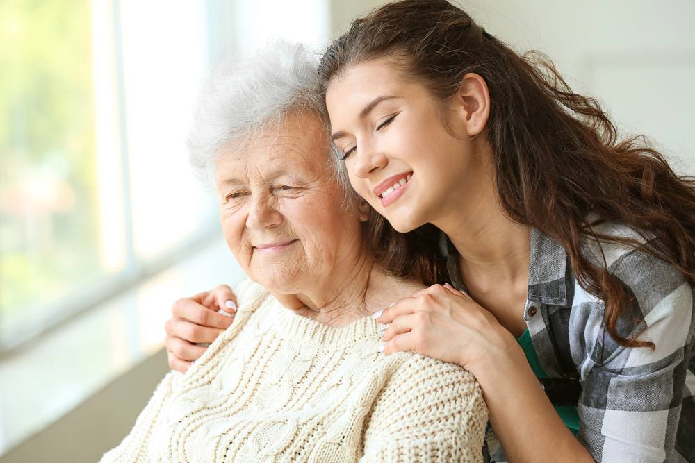 A young woman lovingly hugs an elderly woman, who is smiling and wearing a cream-coloured jumper. They both appear happy and relaxed, with natural light coming through a window in the background.