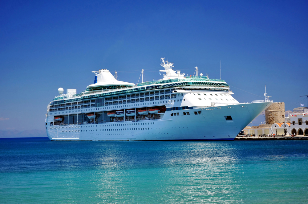 A large white cruise ship is docked at a port on a sunny day, with clear blue sky and turquoise water surrounding it. The ship features multiple decks and lifeboats along the sides.