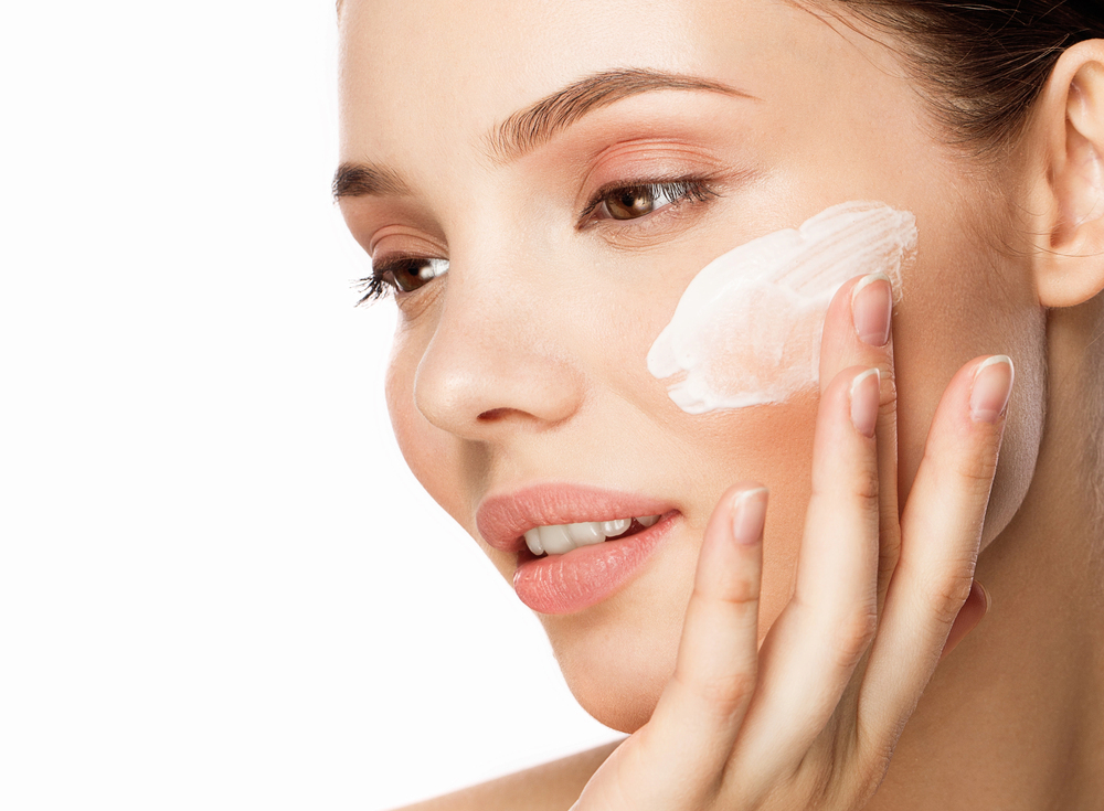 A woman gently applies a dab of white cream to her cheek with her fingers, showcasing smooth, healthy skin against a white background.