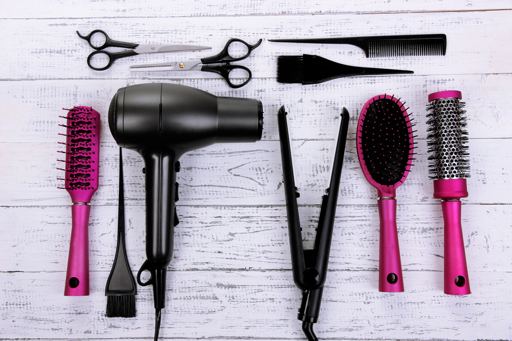 A set of hair styling tools on a white wooden surface, including a hair dryer, straighteners, scissors, combs, brushes, and sectioning clips, with many tools featuring pink handles.