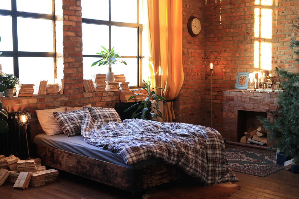 Cosy bedroom with a checked blanket on a rustic wooden bed, exposed brick walls, large windows, potted plants, stacked books, fireplace with firewood, an area rug, and warm sunlight streaming through yellow curtains.