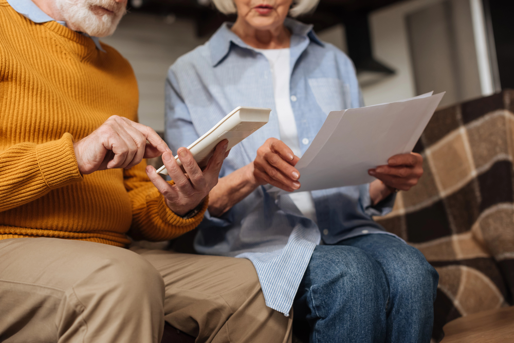 An older couple sits closely together on a sofa. The man, in a mustard jumper, uses a calculator while the woman, in a blue blouse, holds papers. They appear to be discussing finances or bills.