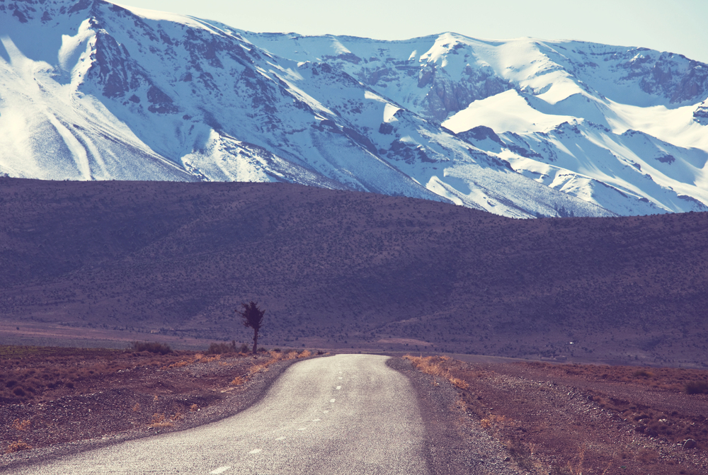 A tarmacked road curves through a barren landscape towards tall mountains with snow-covered peaks under a clear blue sky. Sparse vegetation lines the roadside.