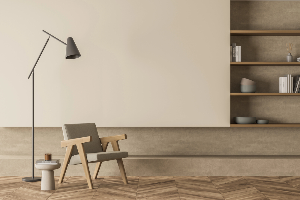 Minimalist living room with a modern armchair, small round side table, floor lamp, built-in wooden shelves with decor items and books, and herringbone wood flooring against neutral beige walls.