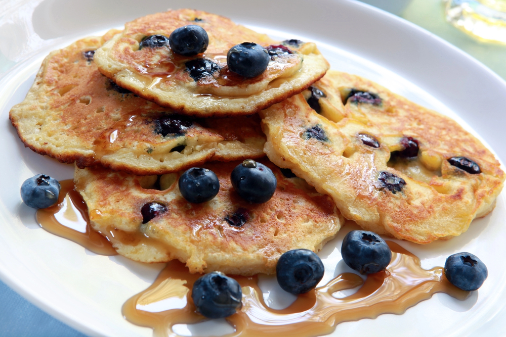 A close-up of a stack of blueberry pancakes on a white plate, topped with fresh blueberries and drizzled with syrup.