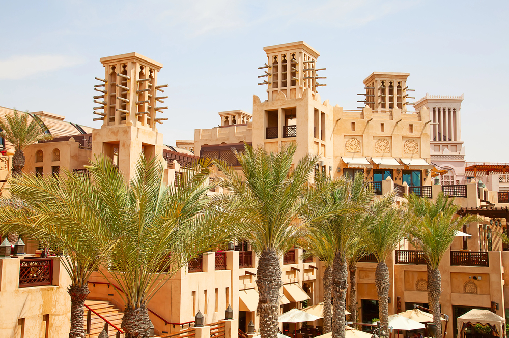 Traditional Middle Eastern architecture with beige wind towers and intricate details, surrounded by tall palm trees under a clear blue sky, creating a warm and inviting atmosphere.