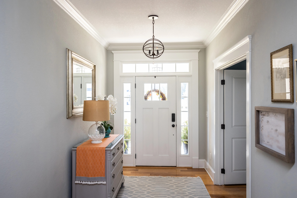 Bright entrance hall with white front door, glass panels, a side table with a lamp, orange runner, mirror, framed art, and light grey walls. Wooden floor and patterned rug complete the welcoming space.