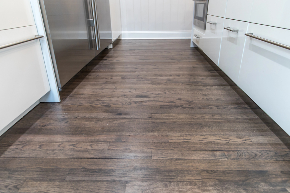 A modern kitchen with dark wooden flooring, white cupboards with long metal handles, a stainless steel fridge, and a built-in oven. The room is well-lit and clean.