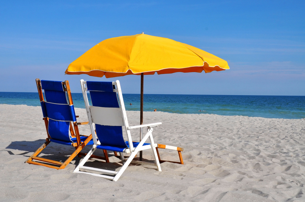 Two blue and white beach chairs sit under a large yellow parasol on a sandy beach, facing the calm blue sea under a clear sky.