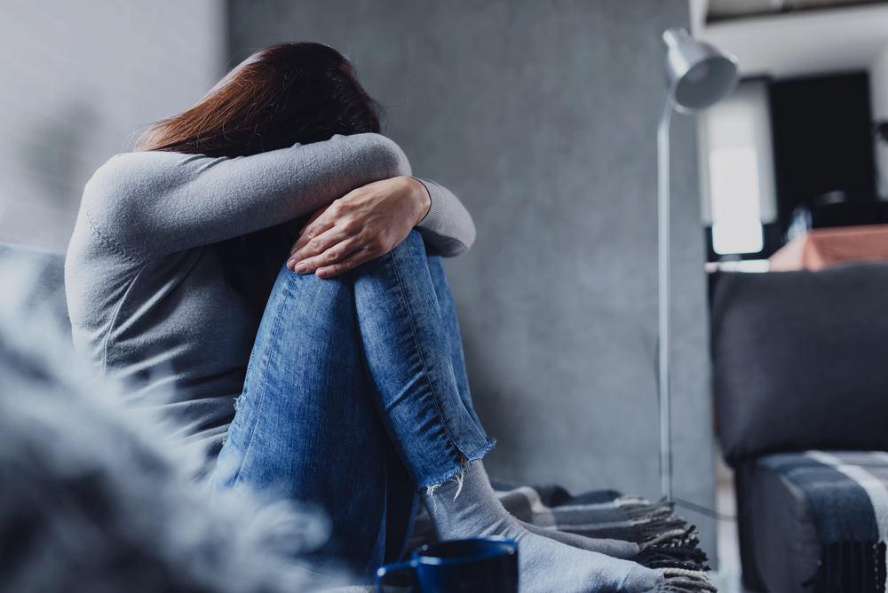 A woman sits on a sofa with her knees drawn up and arms crossed over them, head resting on her arms, appearing distressed or upset in a dimly lit room.