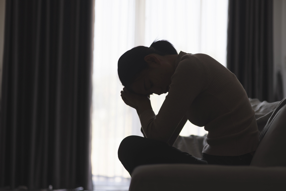 A woman sits on a settee indoors, leaning forward with her elbows on her knees and her hands against her forehead, appearing deep in thought or distressed. She is silhouetted by light coming through large curtains.