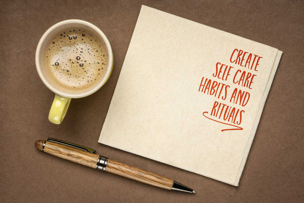 A yellow cup of coffee, a brown and gold pen, and a serviette with the words Create self care habits and rituals written in red are arranged on a brown surface.