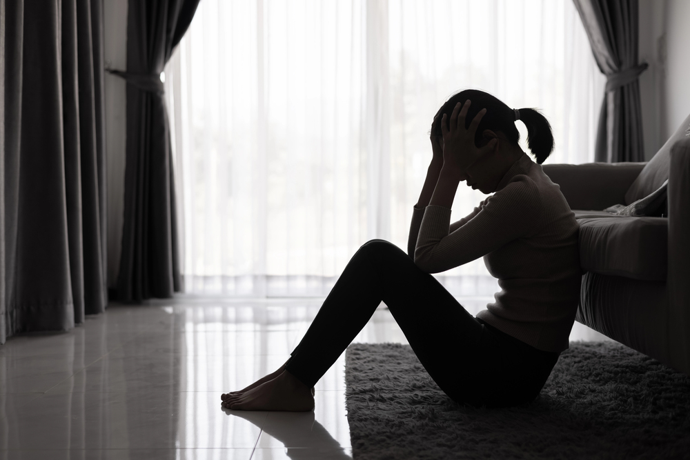 A woman sits on the floor beside a sofa with her knees up and head in her hands, appearing distressed. The room is dimly lit, with large windows and drawn curtains in the background.