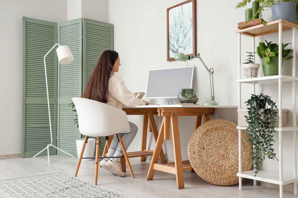 A woman with long dark hair sits at a wooden desk with a computer in a bright, modern home office decorated with green plants and light-coloured furniture.