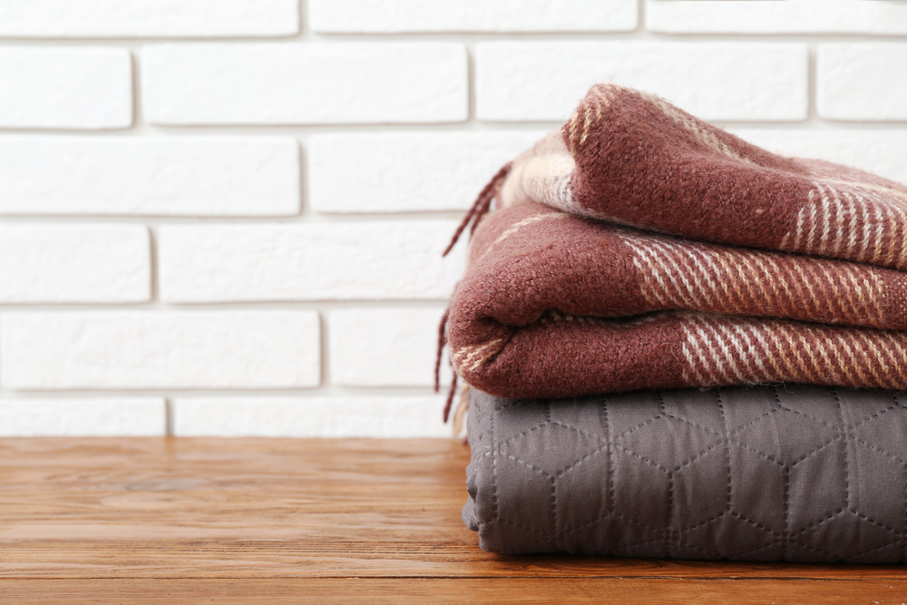 Two folded blankets, one brown checked and one grey quilted, are stacked on a wooden surface with a white brick wall in the background.