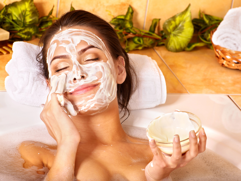 A woman relaxing in a bath applies a creamy facial mask to her face with a towel, smiling with eyes closed. She holds a small bowl of cream, and there are plants and rolled towels in the background.