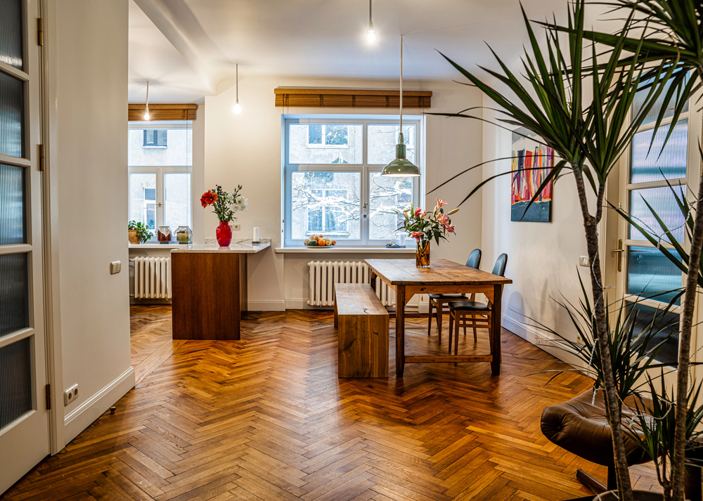 Bright, modern dining area with large windows, wooden herringbone floor, a wooden table with chairs and bench, indoor plants, hanging lights, and decorative items on a side counter.