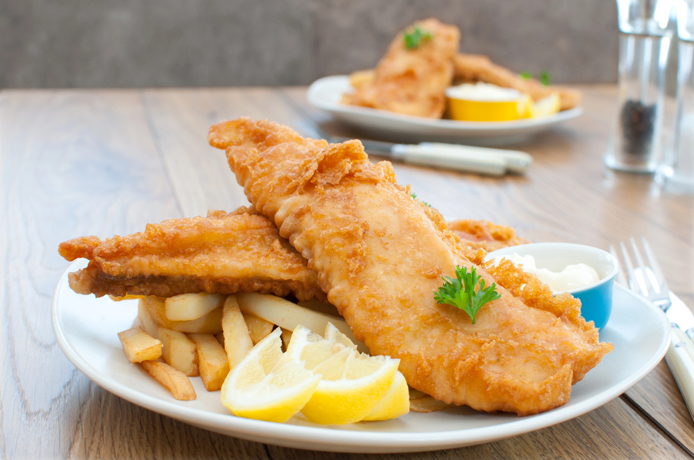 A plate of golden-brown fried fish fillets served with chips, lemon wedges, and a small bowl of tartare sauce. Another similar plate is blurred in the background on a wooden table.