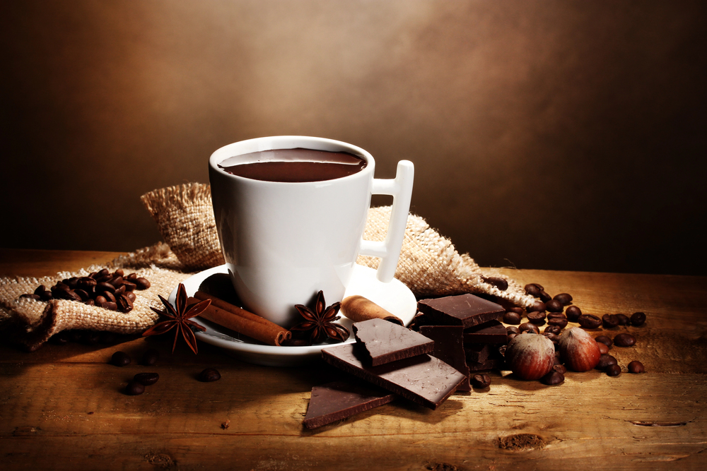 A white mug filled with hot chocolate sits on a saucer, surrounded by pieces of dark chocolate, cinnamon sticks, star anise, coffee beans, hazelnuts, and a hessian sack on a rustic wooden table.