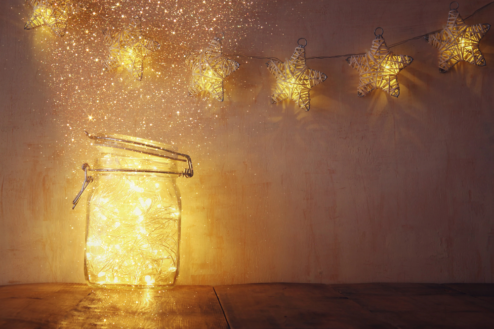 A glass jar filled with glowing fairy lights sits on a wooden surface, with golden sparkles rising from it. Star-shaped string lights hang on the wall in the background, creating a warm, magical atmosphere.