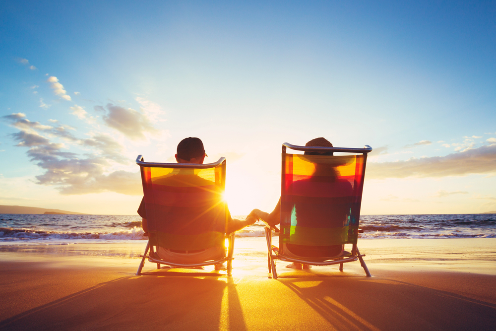 Two people sit in deckchairs on a sandy shore, holding hands and facing the sea as the sun sets, casting a warm golden glow over the water and sky.