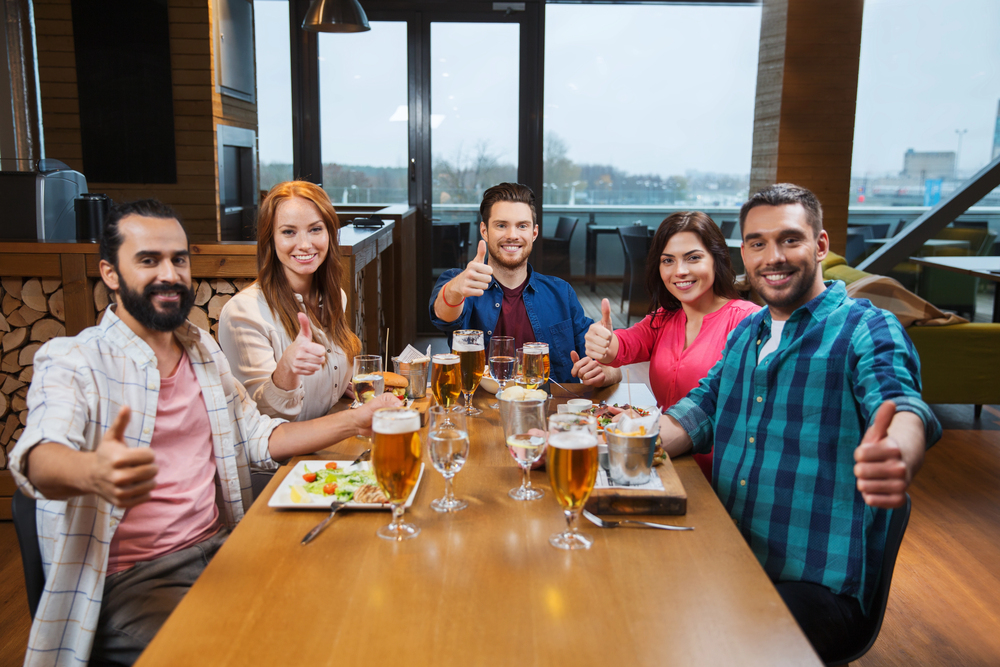 Five smiling people sit at a restaurant table with food and drinks, giving thumbs up and looking at the camera. The setting is bright, modern, and casual with large windows in the background.