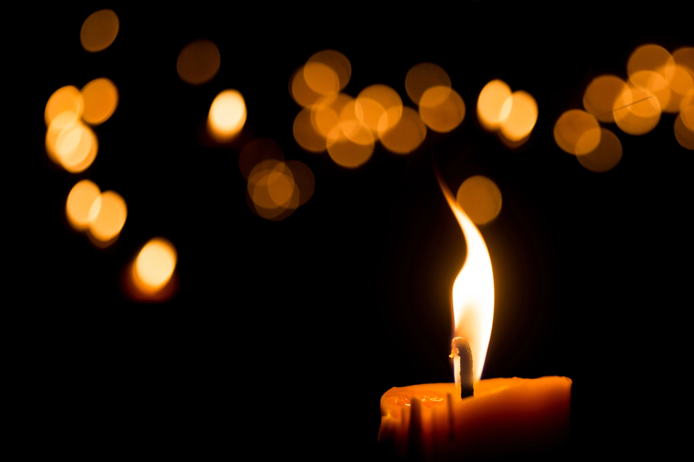 A single lit candle with a bright flame in the foreground, set against a dark background with soft, out-of-focus golden lights creating a warm, glowing effect.