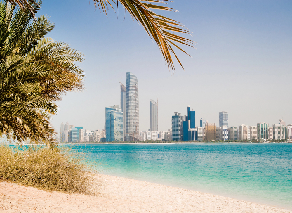 A sandy beach with clear turquoise water is in the foreground, shaded by palm trees, with a modern city skyline of tall glass buildings under a clear blue sky in the background.