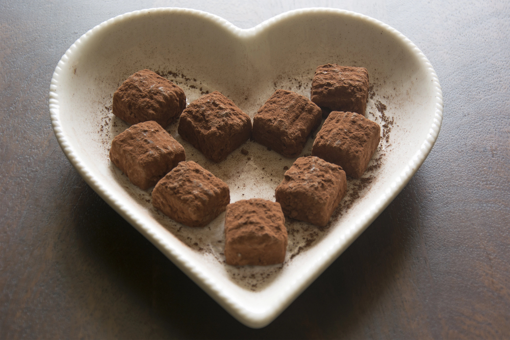 Heart-shaped white dish holding ten square chocolate truffles dusted with cocoa powder, arranged in a heart pattern on a dark wooden surface.