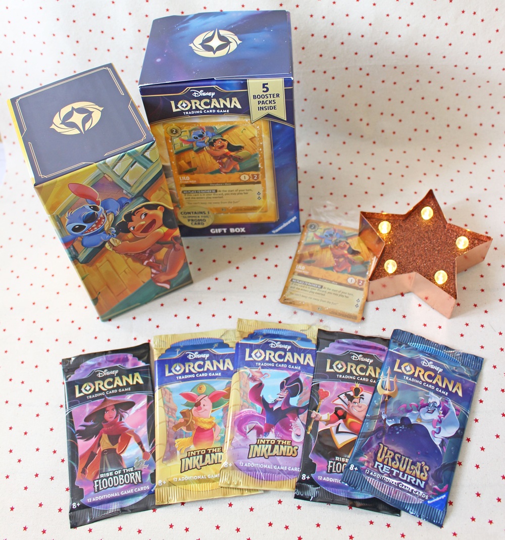 A Disney Lorcana card game gift box, five booster packs, two visible playing cards, a decorative star light, and a character-themed deck box are displayed on a white surface with red polka dots.