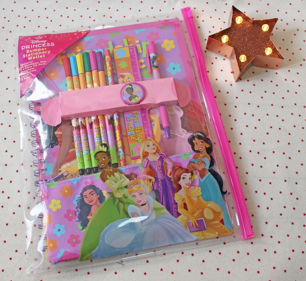 A Disney Princess stationery set in a pink zippered pouch, including pencils, pens, a notebook, and a pencil case featuring various princesses. A lit star-shaped lamp sits beside it on a spotty surface.