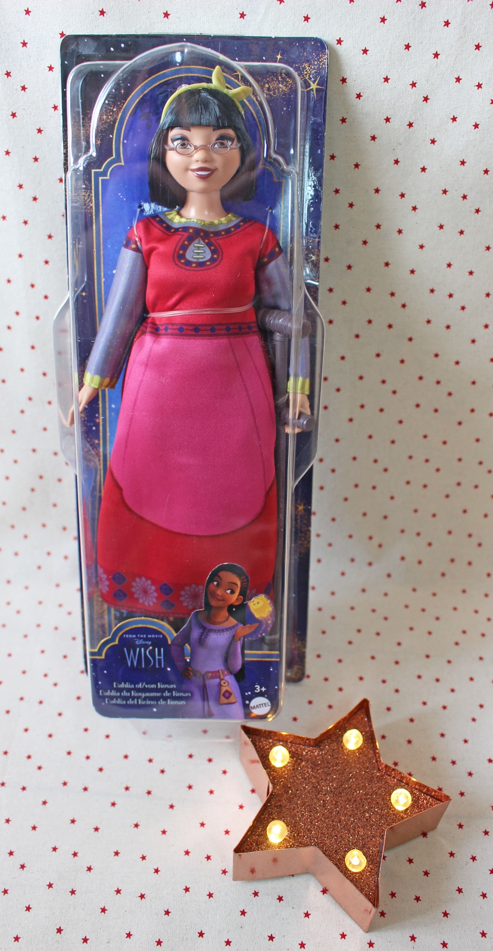 Disney Wish doll in red and pink dress, glasses, and headband, in original packaging with character illustration. In front, a brown star-shaped decoration with small lights. Background is white with red dots.