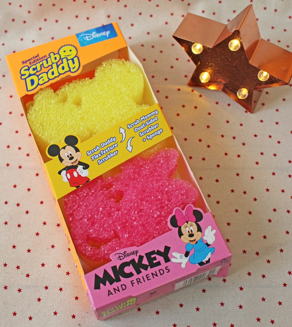 A Scrub Daddy Disney set with yellow and pink sponges shaped like Mickey and Minnie Mouse, in a box decorated with stars and cartoon images of Mickey and Minnie. A lit star-shaped lamp is beside the box on a star-patterned surface.