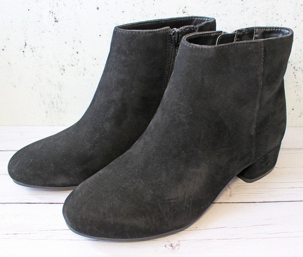 A pair of black suede ankle boots with low heels and side zips, placed on a light wooden surface against a white background.