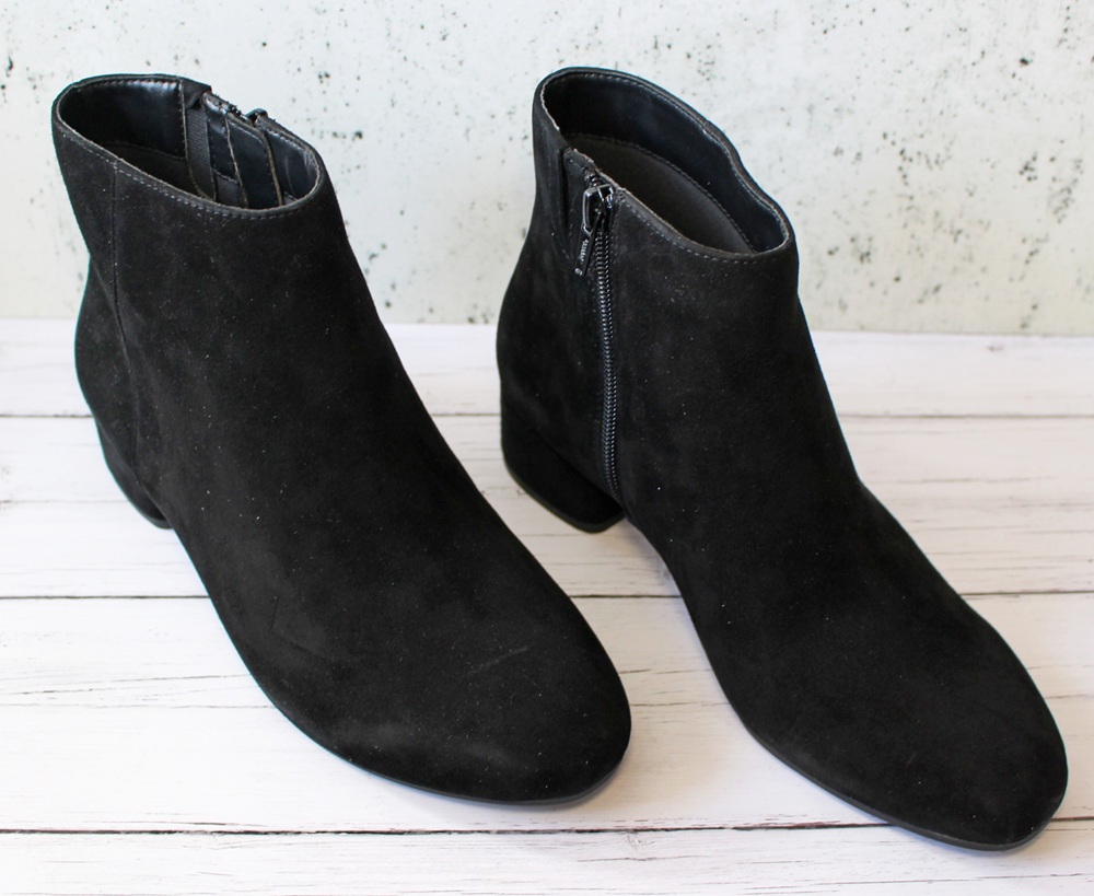 A pair of black suede ankle boots with side zips, placed on a white wooden surface against a lightly speckled background.