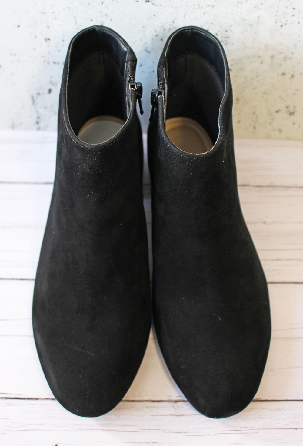 A pair of black suede ankle boots with side zips, viewed from above, placed on a white wooden surface with a light textured background.