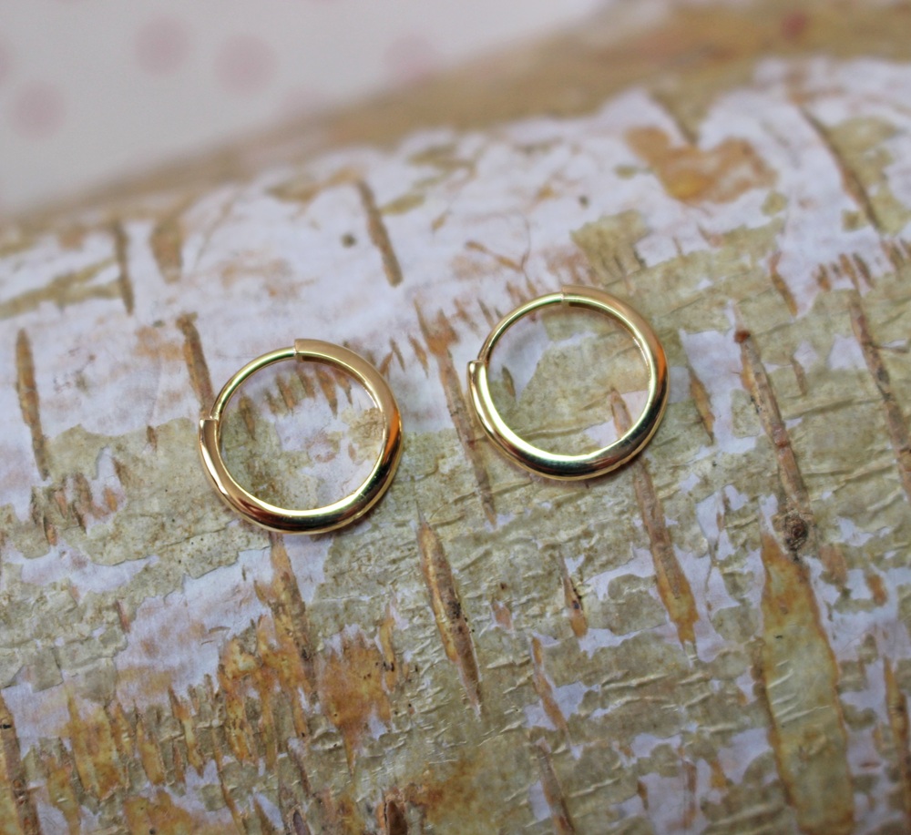 Two small gold hoop earrings are placed side by side on a light-coloured, textured wood surface with natural markings.