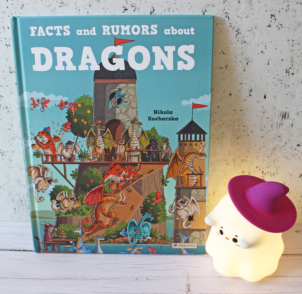 A colourful children’s book titled Facts and Rumours about Dragons with illustrated dragons on the cover stands next to a small, glowing ghost-shaped lamp with a purple hat.