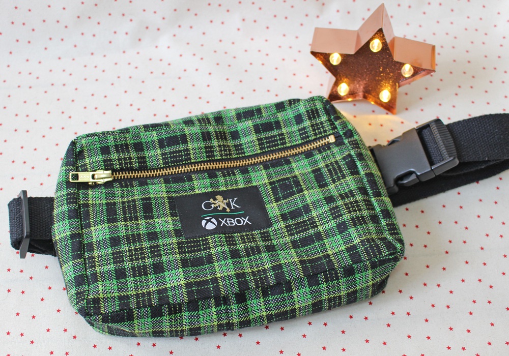 A green and black checked Xbox-branded bum bag with a gold zip is displayed on a white surface with red dots. A lit, brown star-shaped decoration is in the background.