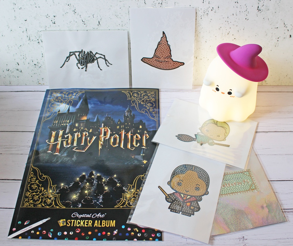 A Harry Potter sticker album, three pixel art character stickers, spider and hat stickers, and a cute white ghost-shaped lamp with a purple hat are arranged on a white wooden surface.