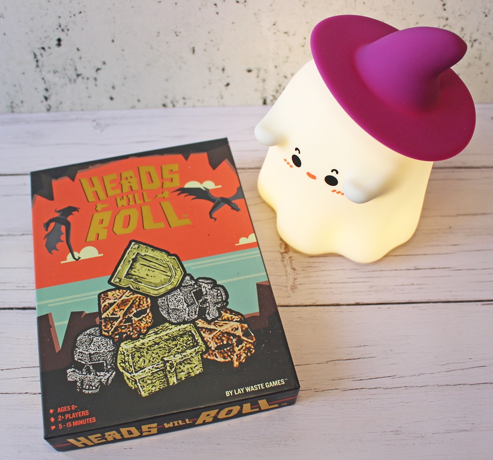 A box of the game Heads Will Roll with skull and treasure illustrations sits on a white wooden surface next to a cute ghost-shaped lamp wearing a purple witch’s hat.