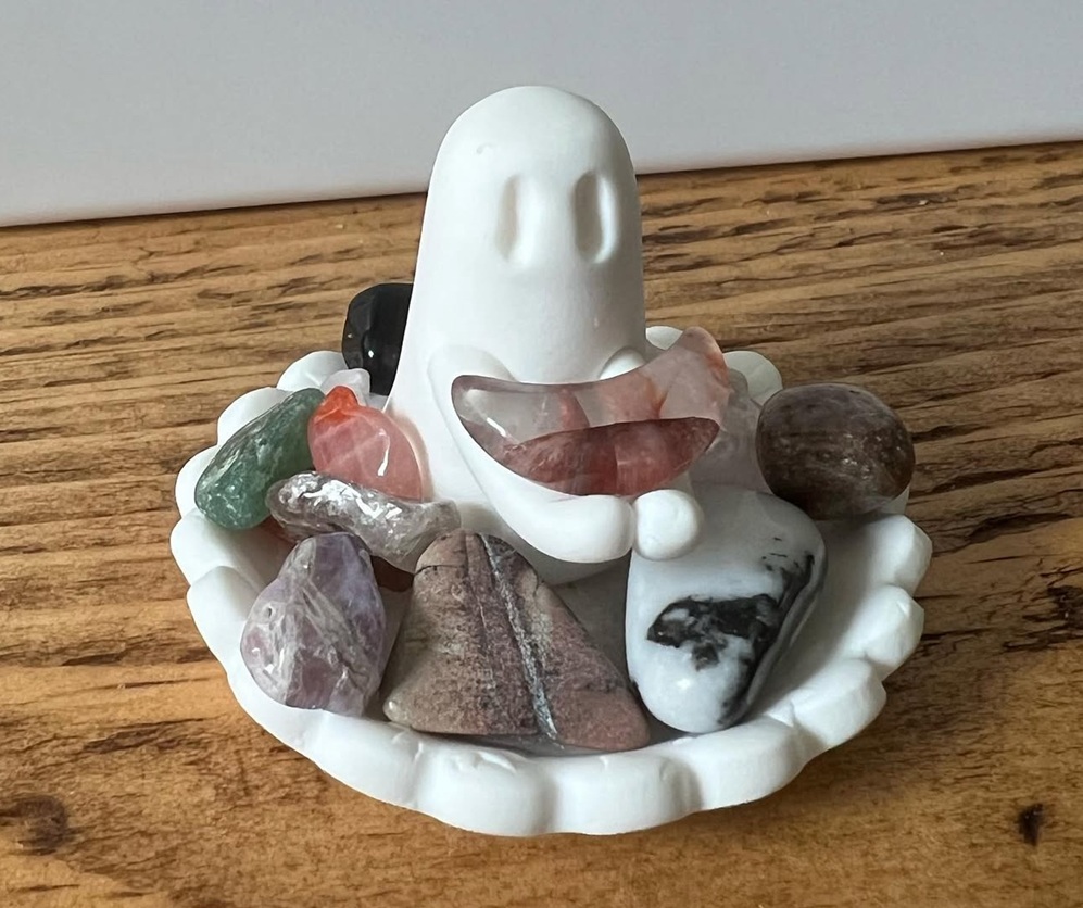 A small white ceramic ghost figurine sits in a scalloped dish, surrounded by various polished crystals and stones, on a wooden surface.