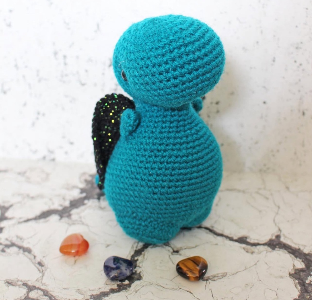 A teal crocheted dragon with black glittery wings stands on a marbled surface, facing away. Three polished stones—orange, purple, and brown—are placed in front of it. The background is a textured white wall.