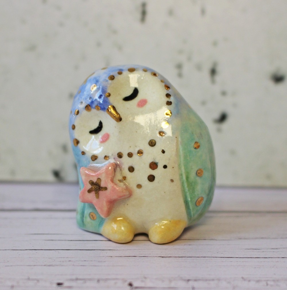 A small ceramic owl figurine with closed eyes, blue and gold spots on its head, green wings, and yellow feet, holding a pink star-shaped object, sitting on a light wooden surface with a speckled background.