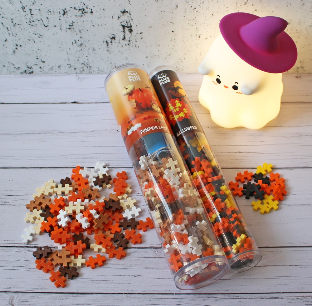 Two clear tubes filled with Plus-Plus puzzle pieces in autumn colours sit on a white wooden surface; some pieces are scattered outside. A cute ghost night light with a pink witch’s hat glows in the background.